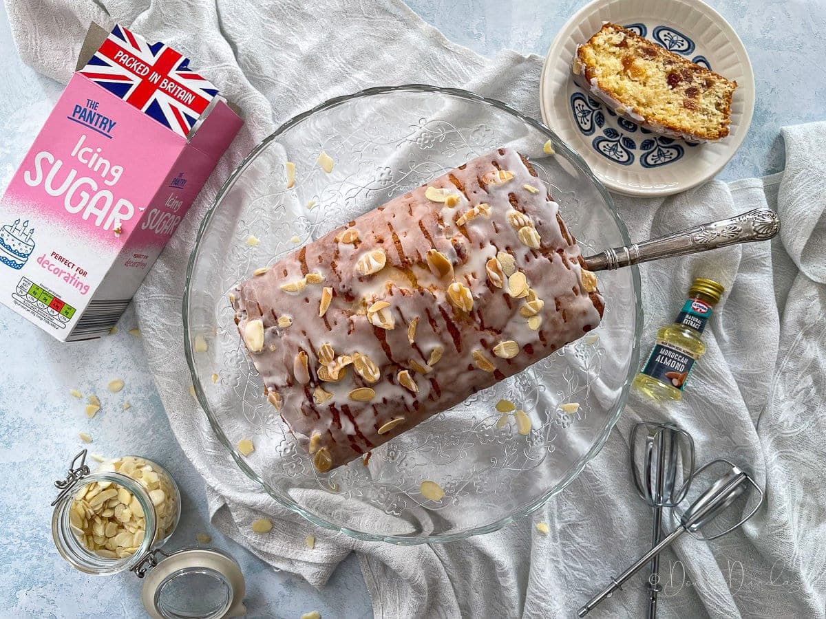 loaf cake topped with icing and toasted almonds on a glass cake stand with a silver slice. Slice of cake on plate to the side.