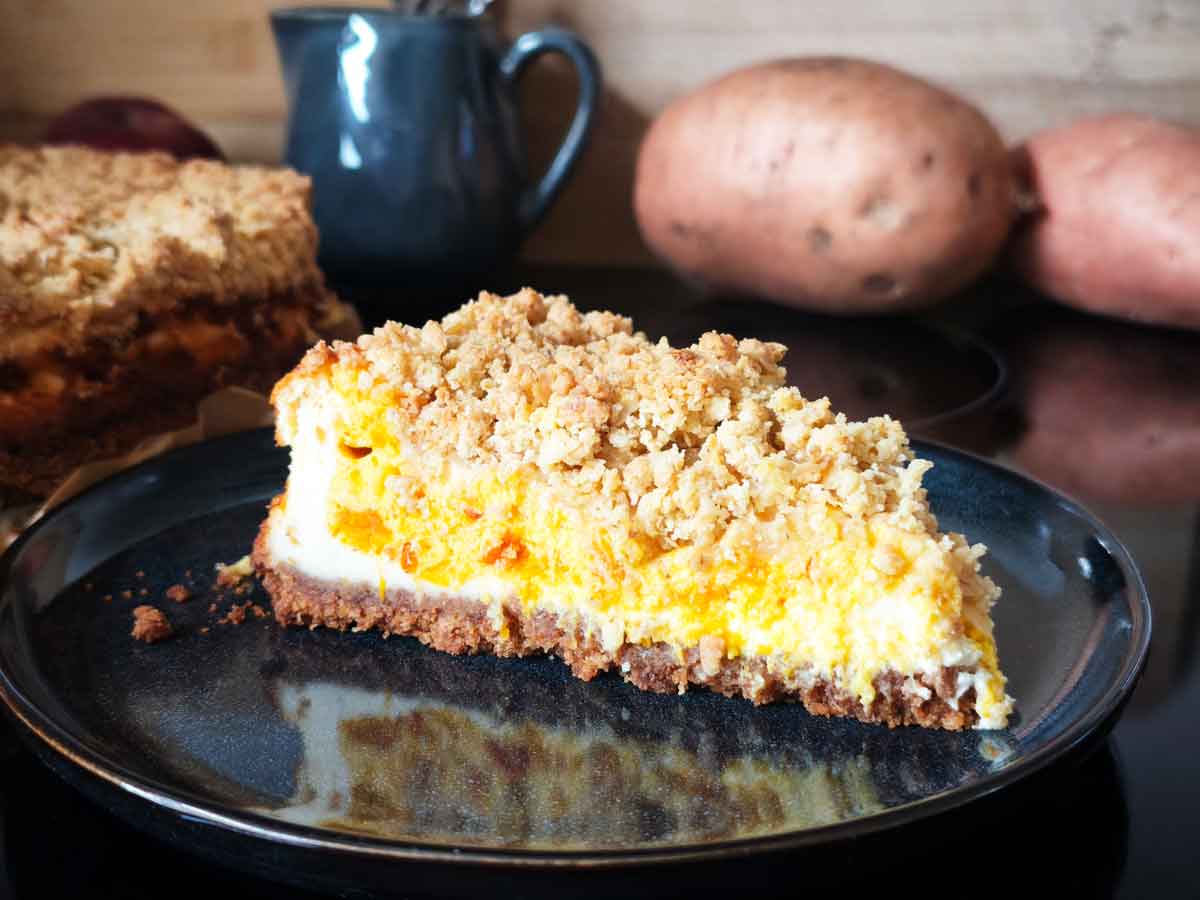 Ein Stück fertiger Pumpkin-Cheese-Pie auf einem Teller. Das Kuchenstück hat einen dunklen dünnen Keksboden, eine Quark-Kürbis-Füllung und dicke Streusel.