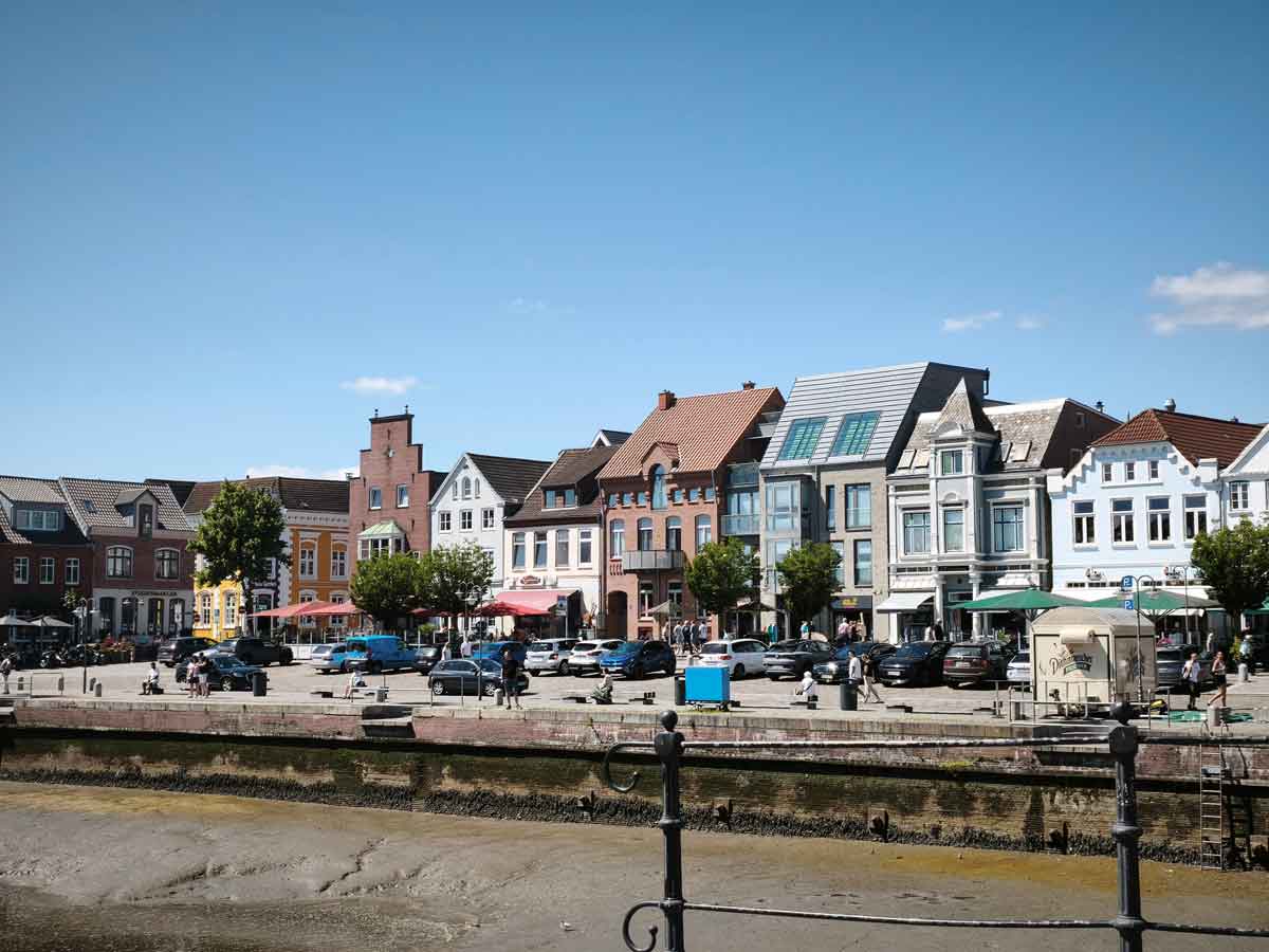Blick auf den Husumer Hafen bei Ebbe. Der Blick ist auf die Hafenhäuser gerichtet, die Cafés, Restaurants und Geschäfte beherbergen. Fußgänger gehen spazieren.
