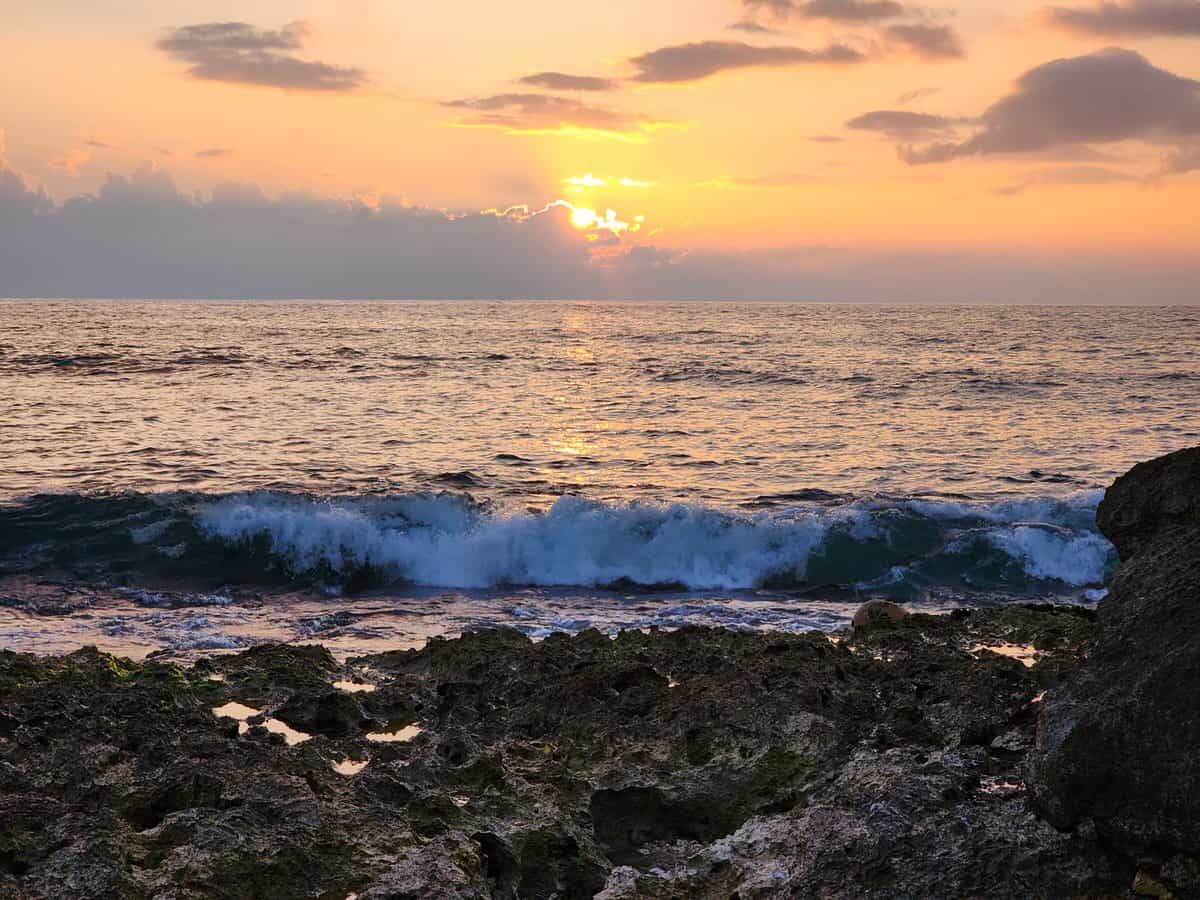 A vibrant sunset over the ocean, with gentle waves crashing against a rocky shoreline. The sky is painted in shades of orange, pink, and purple, creating a serene and picturesque coastal scene.
