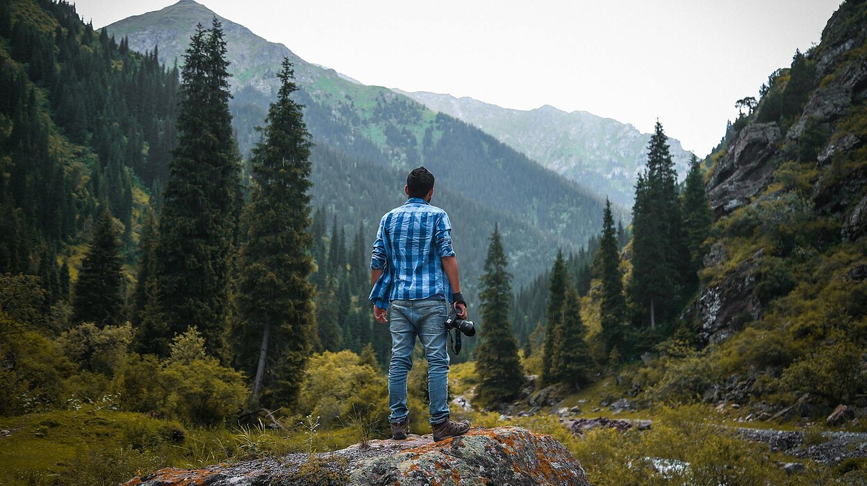 man in blue and white checked quarter sleeved shirt holding black camera standing on rock