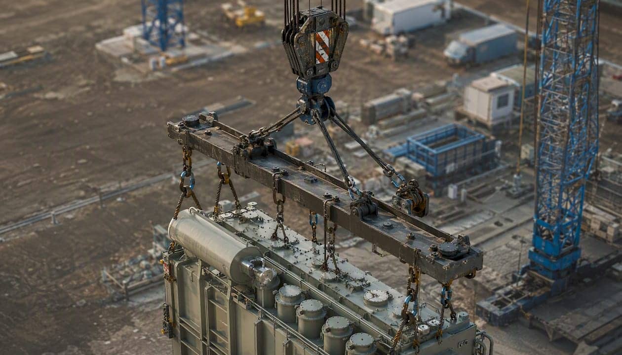 Ein Kran hebt mit einer Traverse einen industriellen Transformator an.