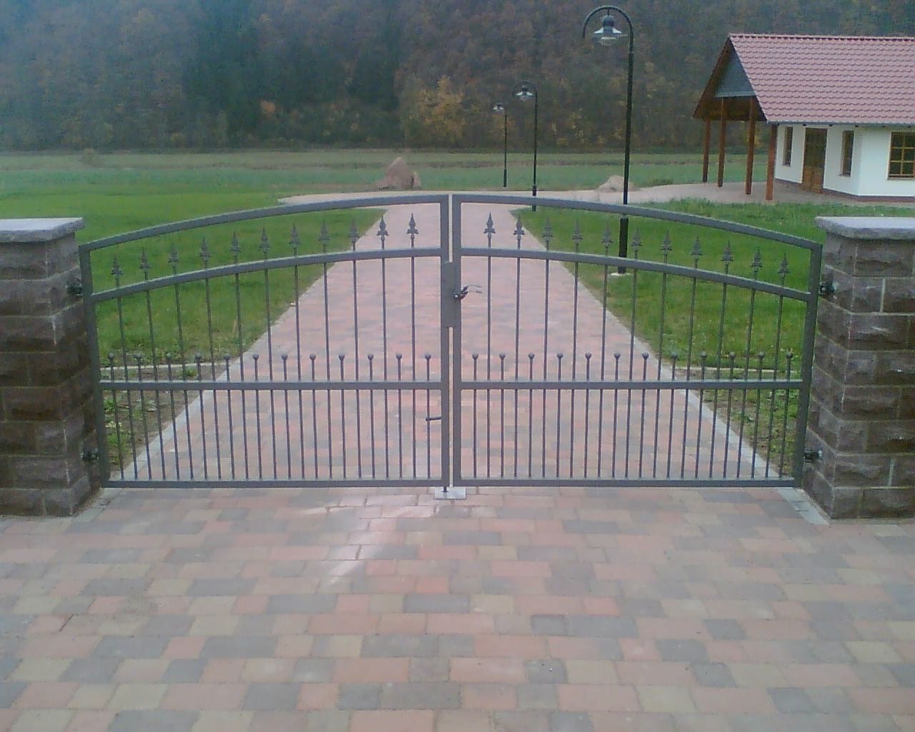 Doppelflügeliges Schmiedeeisentor in Grau mit geschwungenem Oberteil und Zierspitzen, installiert zwischen Steinpfeilern auf einem gepflasterten Zufahrtsweg. Ein Produkt von Tore & Torsysteme von Stahl- und Metallbau Drost.