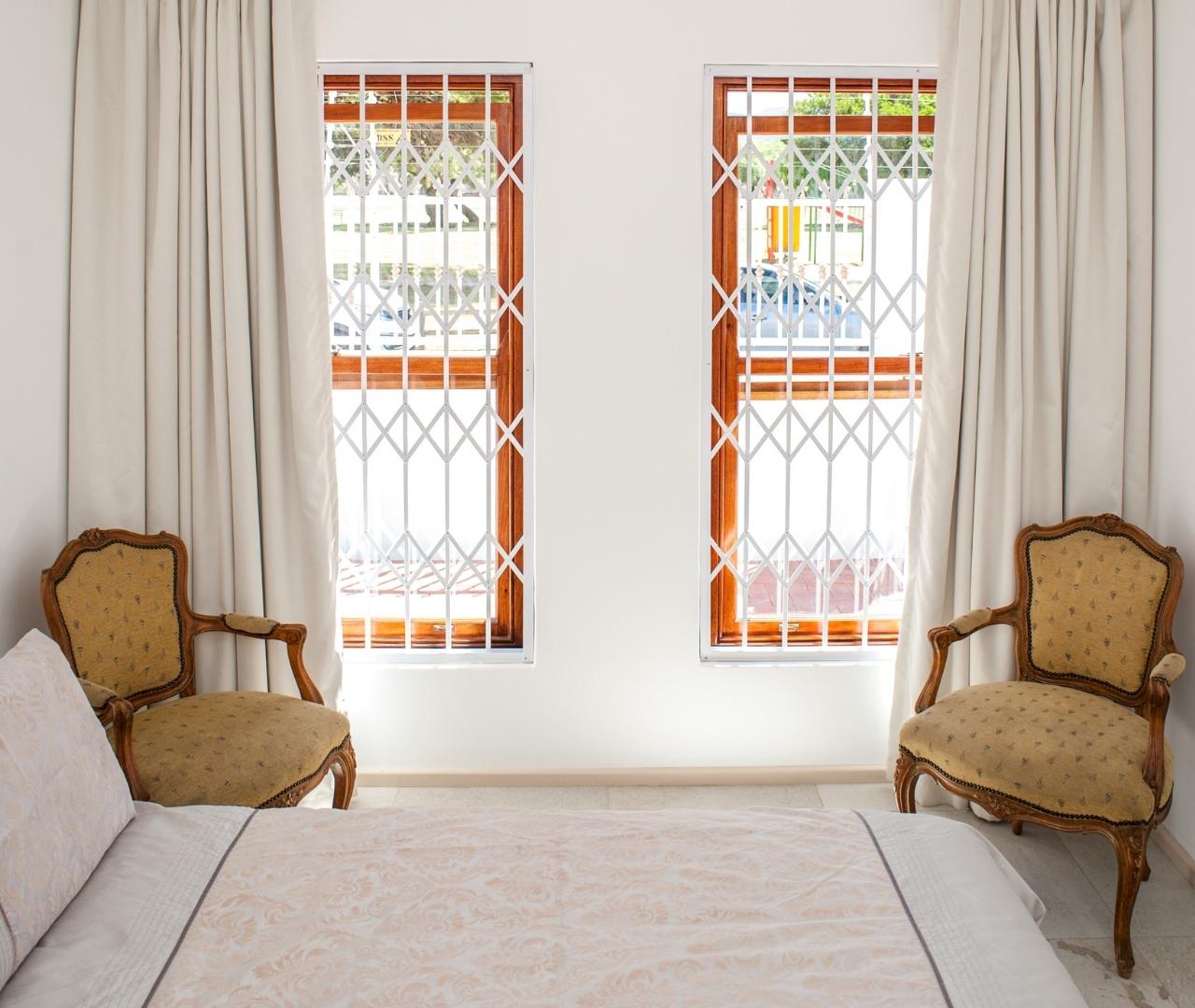 White Magnagrille trellis burglar bars installed on two wooden-framed bedroom windows between classic armchairs.