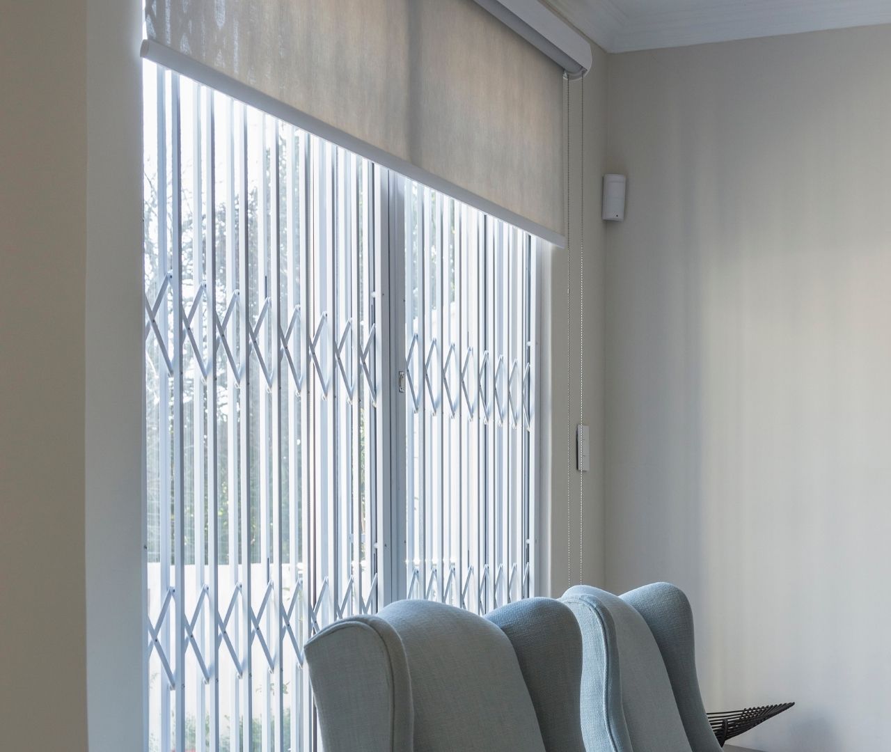 Side view of retractable white burglar door behind roller blind, protecting lounge window next to armchairs.