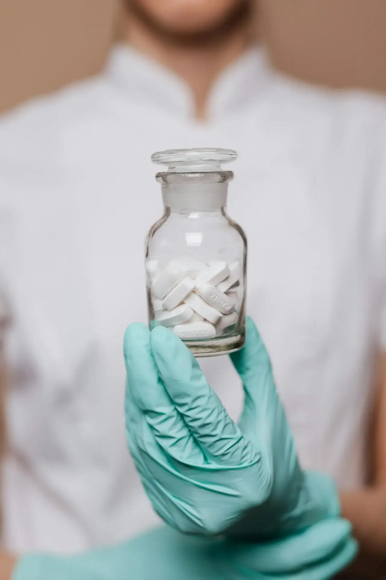 Hand in Handschuhen hält ein Apothekerglas mit Tabletten.