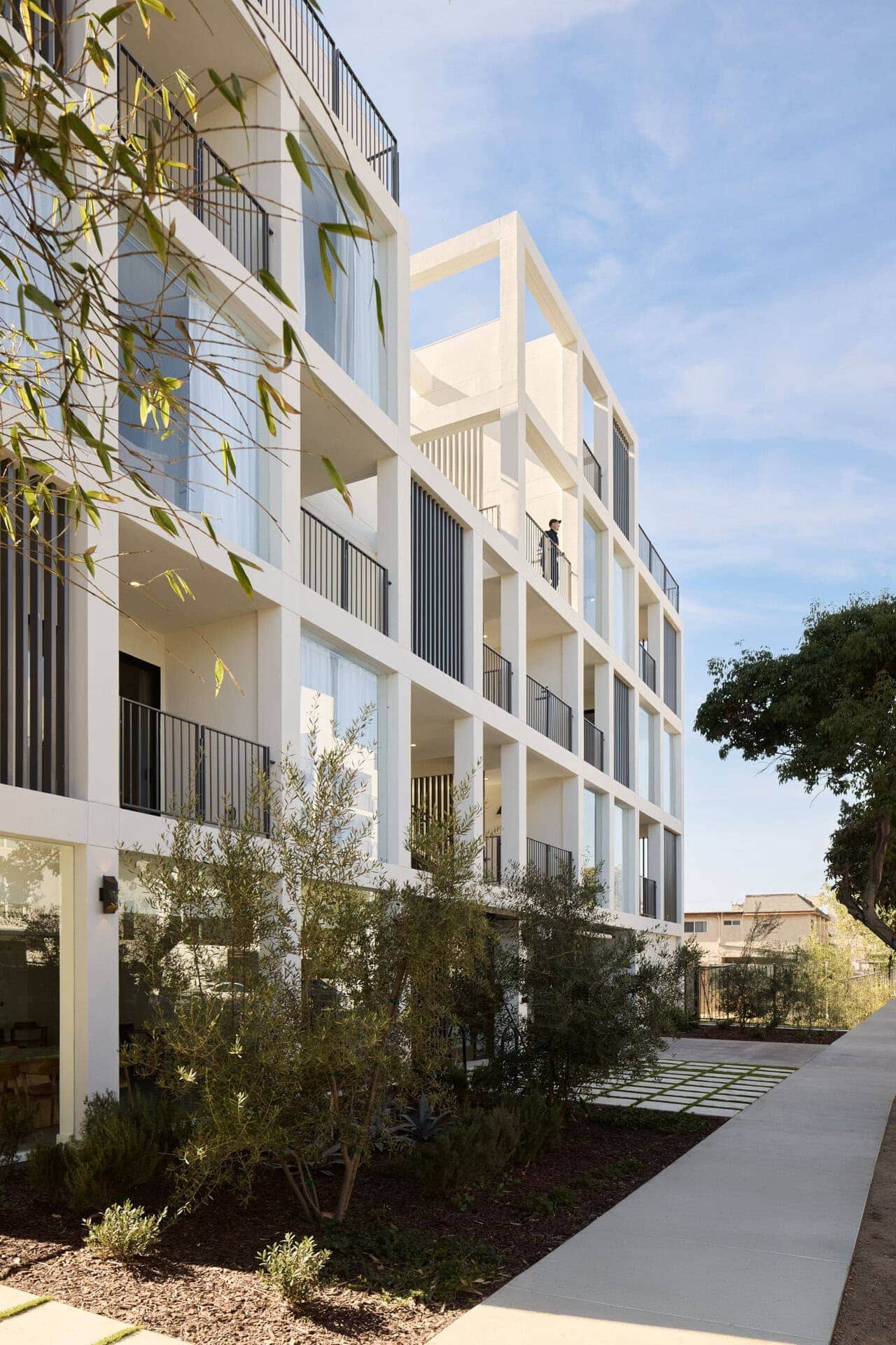 Modern white residential building with open balconies, black metal railings, and large glass windows under a clear blue sky.