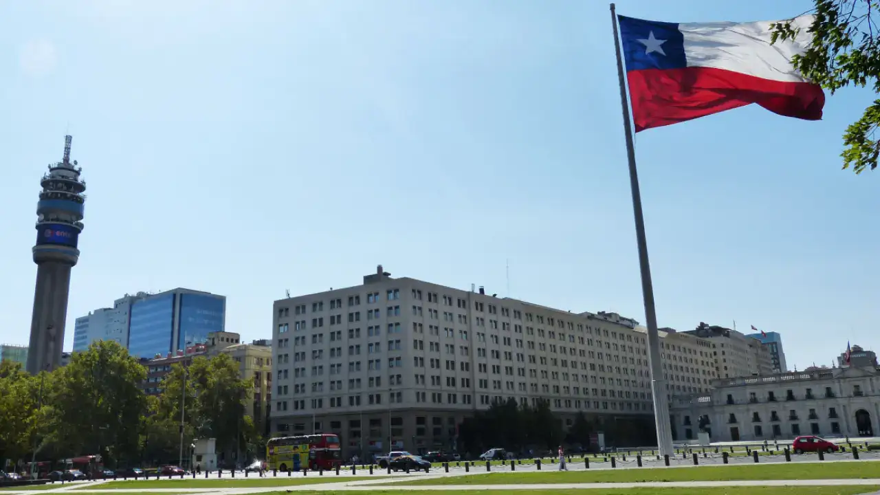 Bandeira do Chile tremulando em destaque, com um edifício histórico ao fundo em Santiago.
