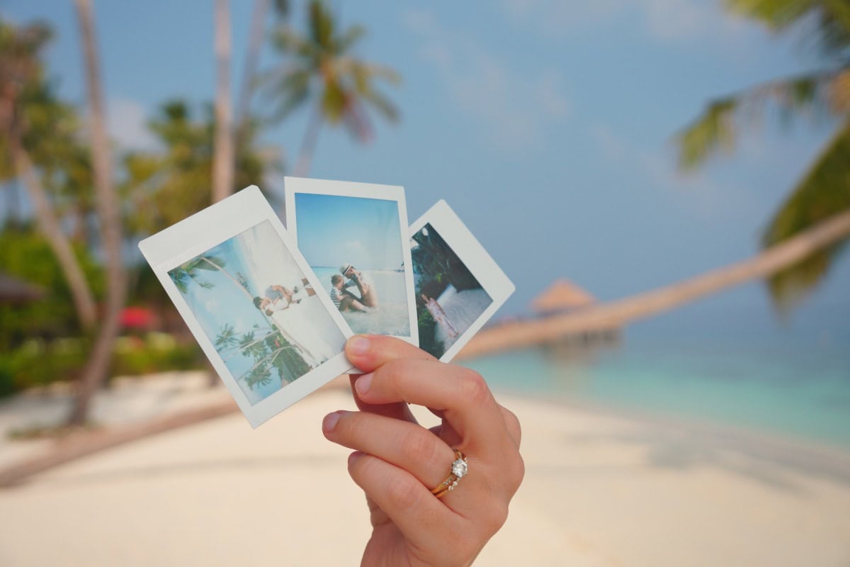 A hand displaying three instant photos of reethi faru resort in the maldives, with a tropical beach on the background