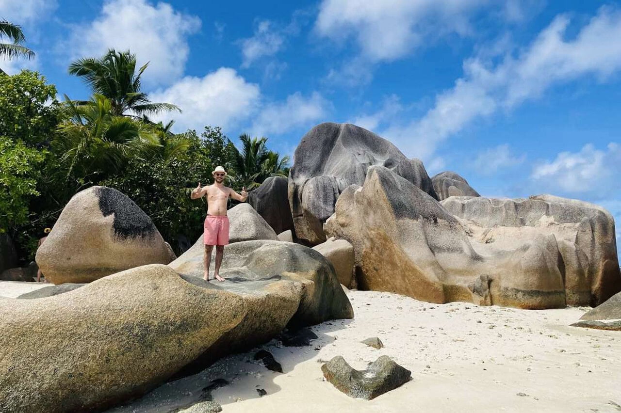 Man standing on giant granite boulders, with lush palms in the background, in Anse Source D'Argent, the most iconic beach in La Digue