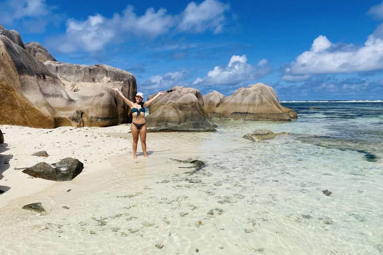 Woman standing in calm turquoise sea water at Anse Source D'Argent beach on a sunny day, with a background of giant granite boulders