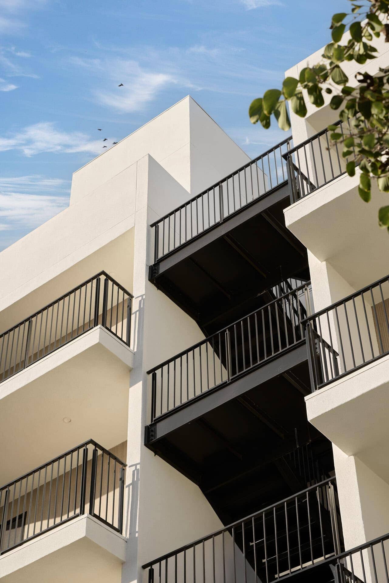 Modern apartment building exterior with black metal fire escape and white stucco walls under a blue sky.