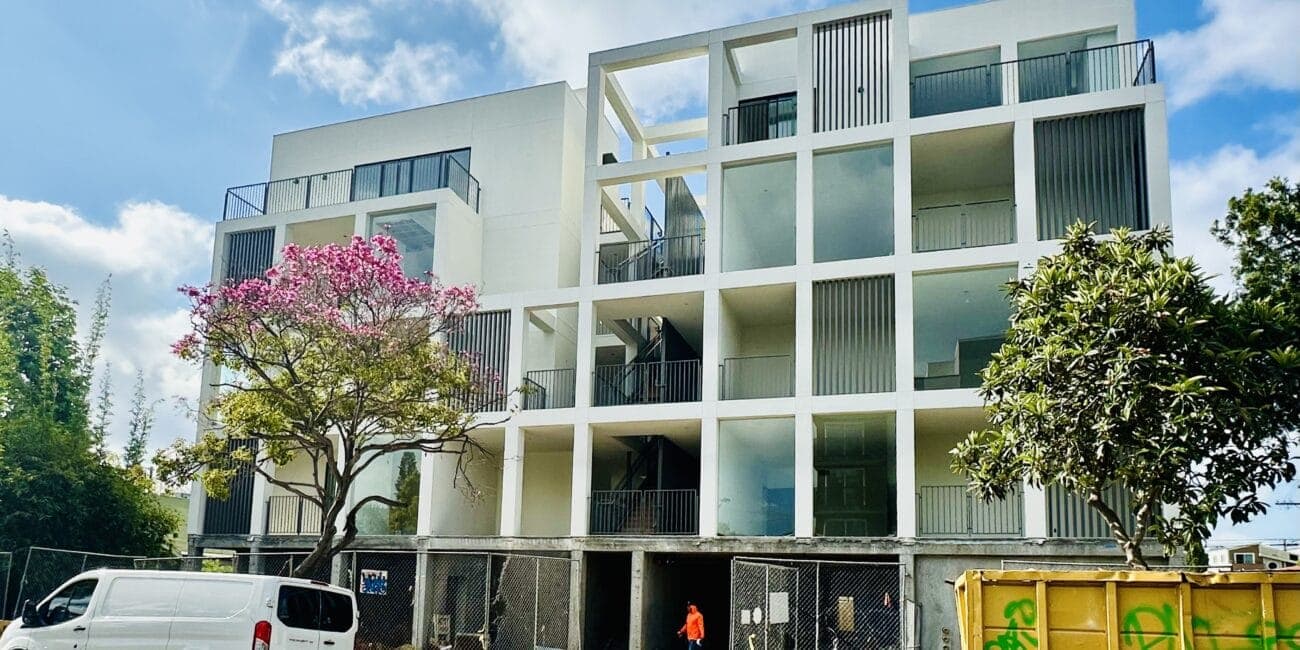Modern white multi-story apartment building with open-air corridors and balconies, featuring vertical slat accents and a geometric facade.