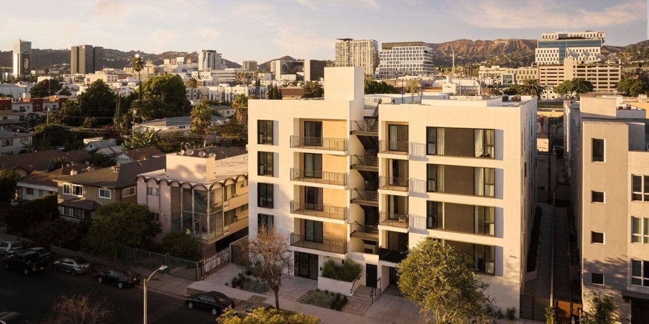 Modern white apartment building with balconies and large windows, set in an urban neighborhood at sunset.