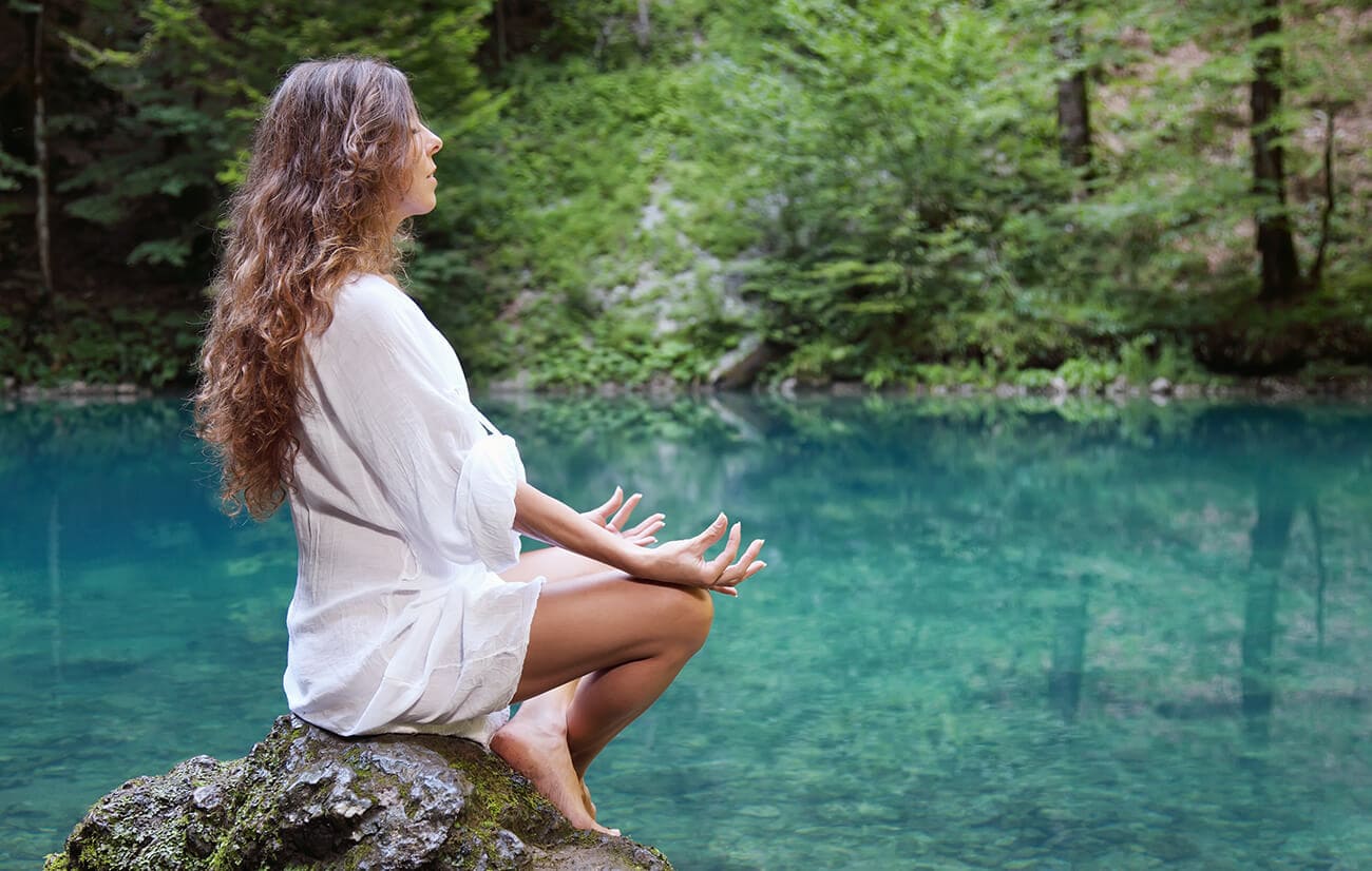 Spiritual Wellness-A woman with long curly hair sits in a meditative pose on a mossy rock beside a calm, turquoise lake surrounded by a lush green forest.