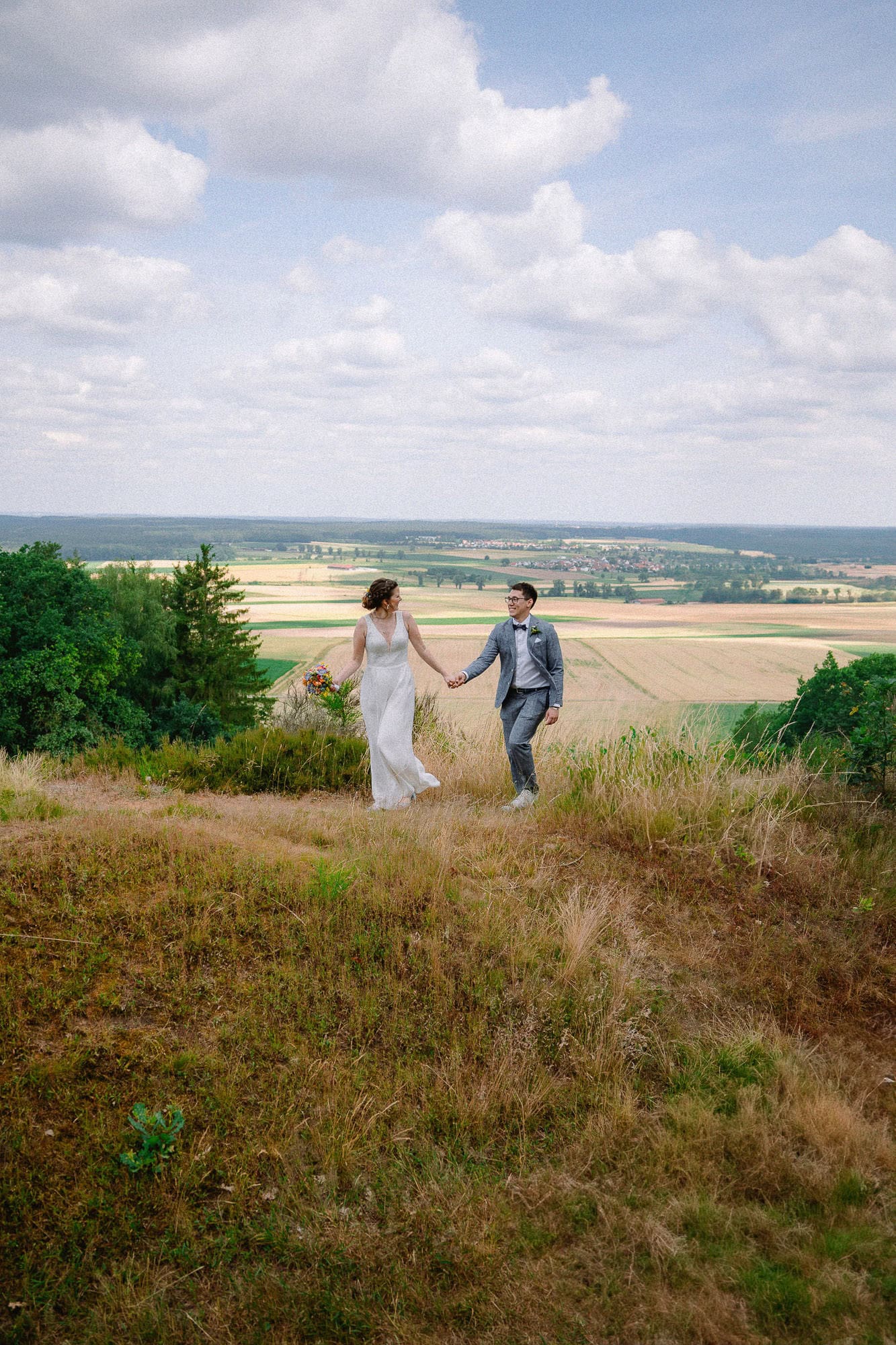 Ein Paar im Hochzeitskleid geht Hand in Hand über einen grasbedeckten Hügel, wunderschön festgehalten von ihrem Hochzeitsfotografen Regensburg. Die Braut hält einen Blumenstrauß, während sich im Hintergrund die malerische Aussicht auf Felder und einen bewölkten Himmel elegant ausbreitet.