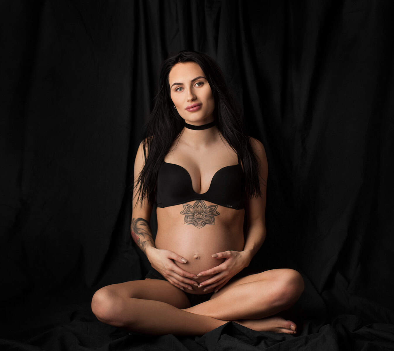 Seksig gravidfotografi av en gravid kvinne med tatoveringer som sitter korslagt foran en svart bakgrunn.