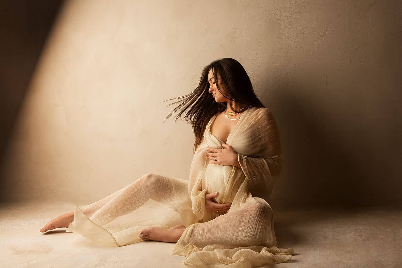 Nydelig gravidfoto av en gravide kvinne som sitter på gulvet med hender på magen, i mykt og varmt lys, perfekt for gravidfotografering og minnerikes bilder.