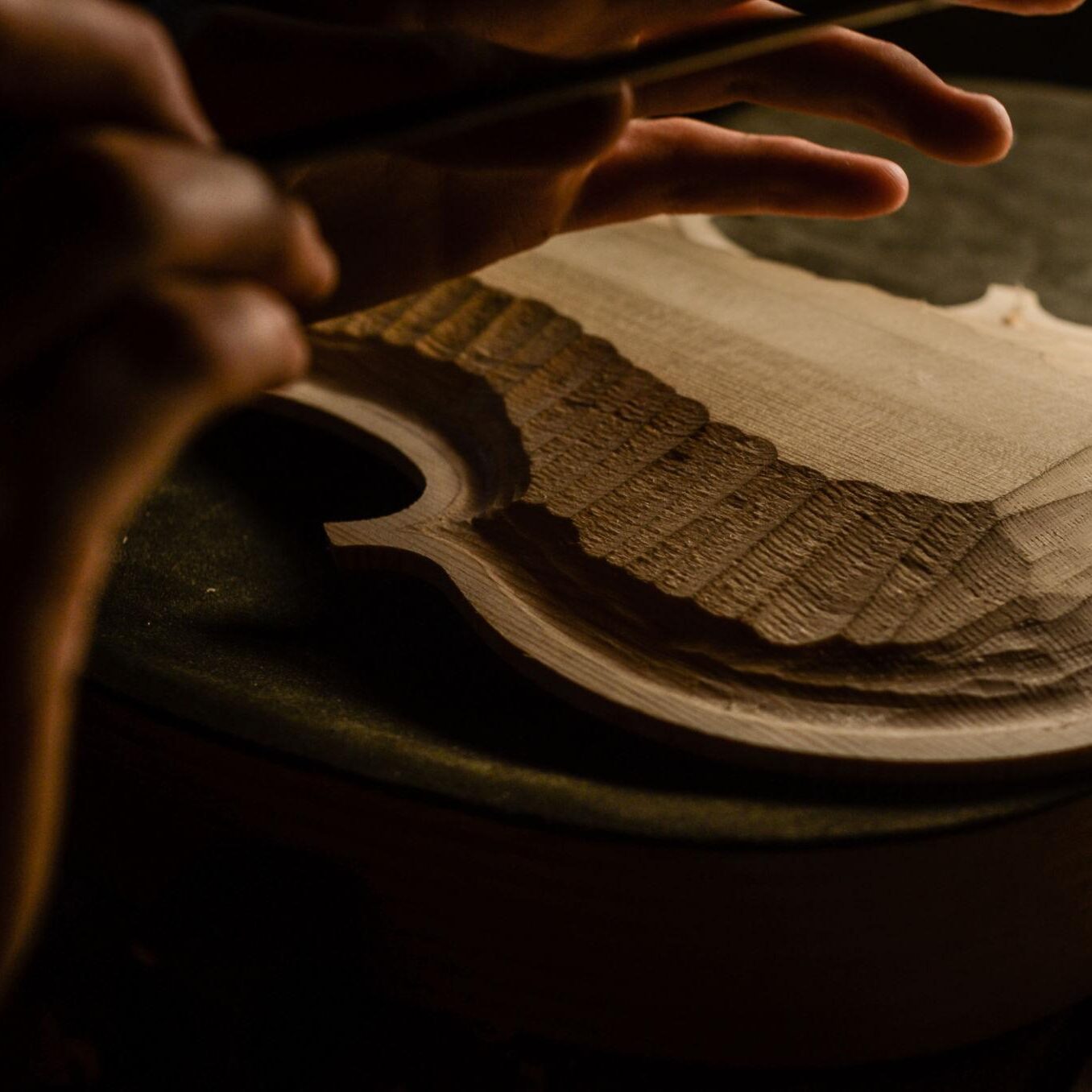 Close-up of hands shaping a wooden shoe sole with a rasp, carving its tread pattern under warm light.