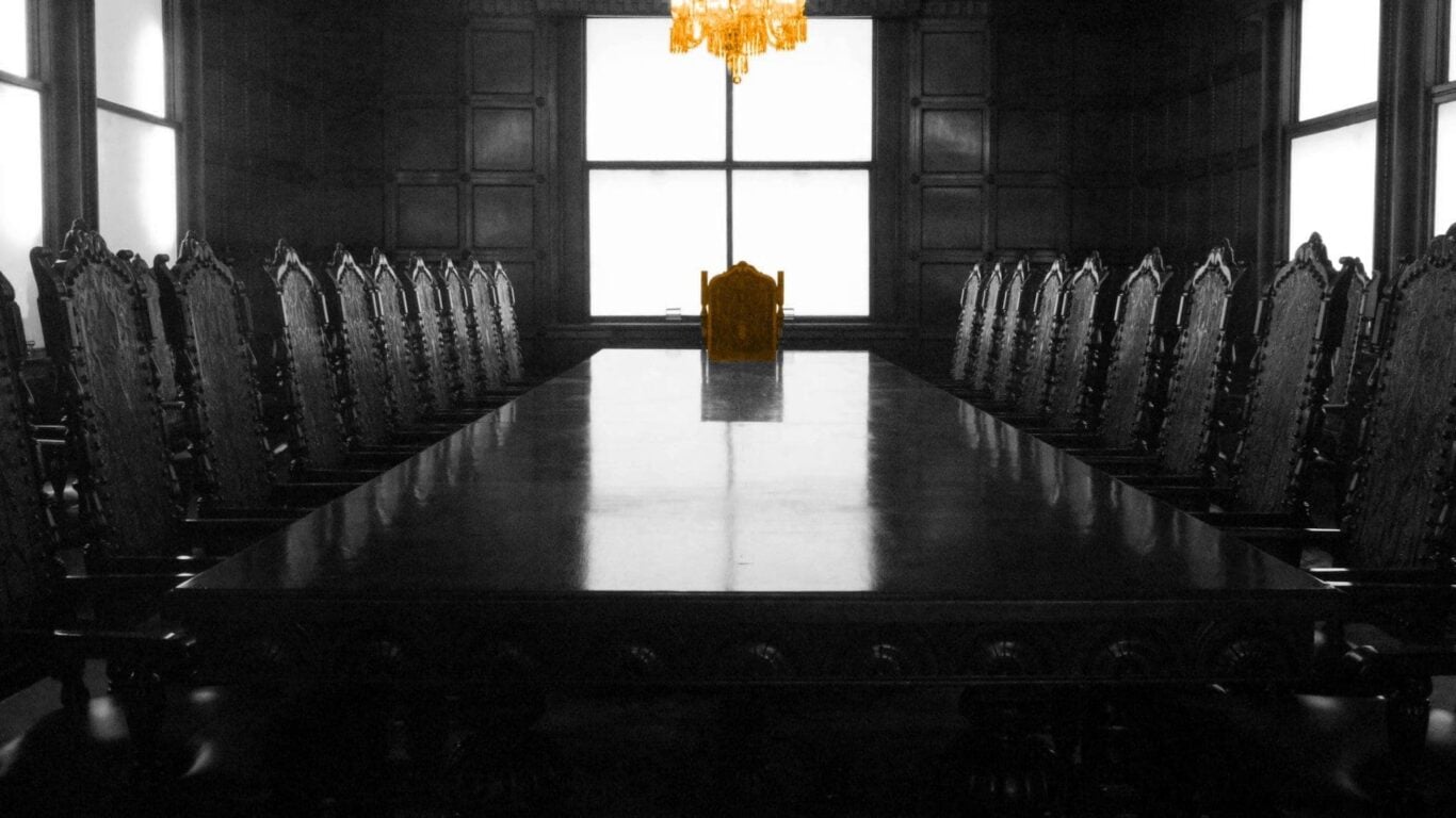 A black and white image of a table with multiple chairs, the chair at the head of the table is orange