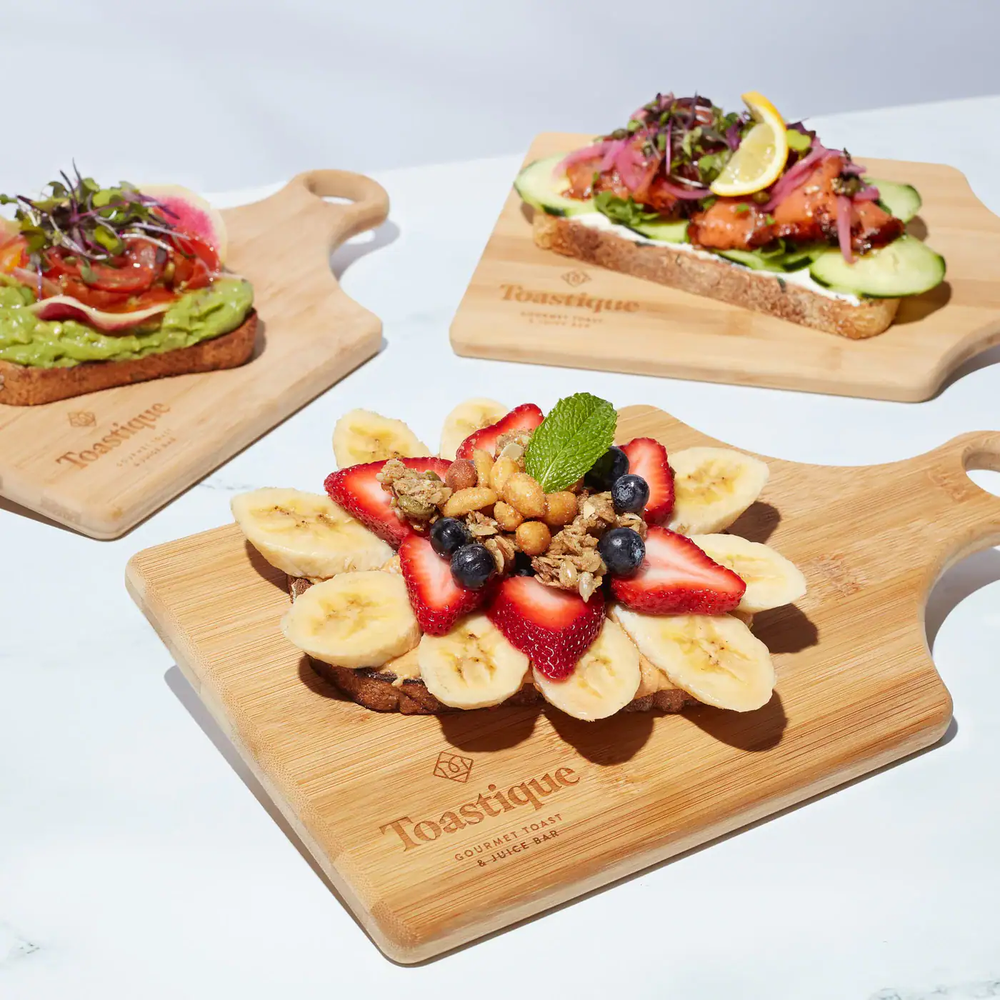 Assorted gourmet toasts served on wooden boards, featuring fruit, avocado, and smoked salmon toppings on a bright white table — the kind of fast, healthy lunch available at Toastique's Raleigh cafe and coffee shop