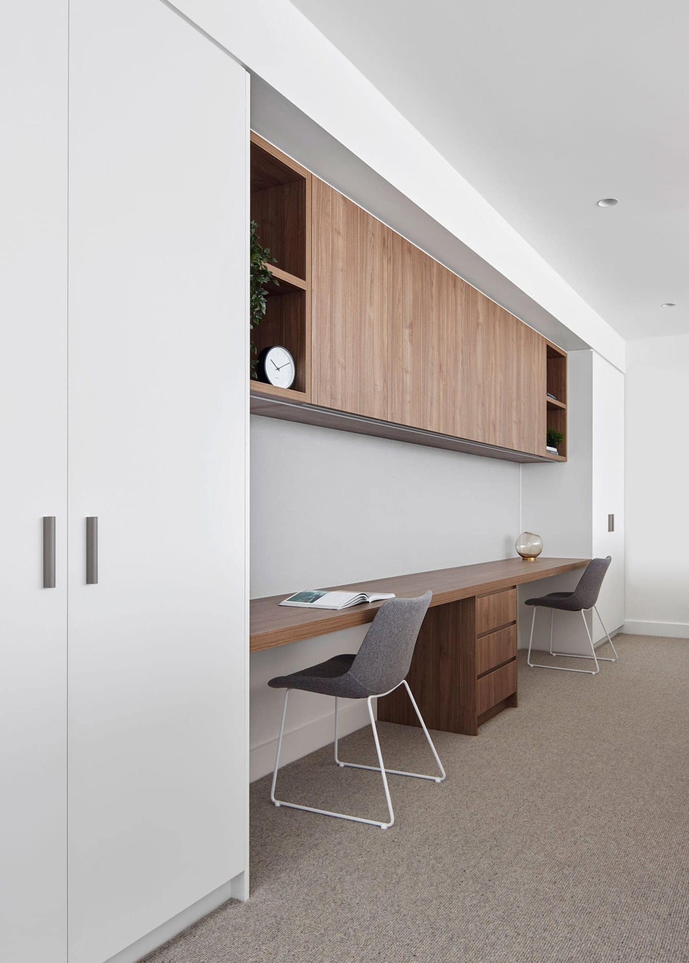 Built-in study nook with a long timber desk, overhead cabinets, white storage, and two grey chairs.