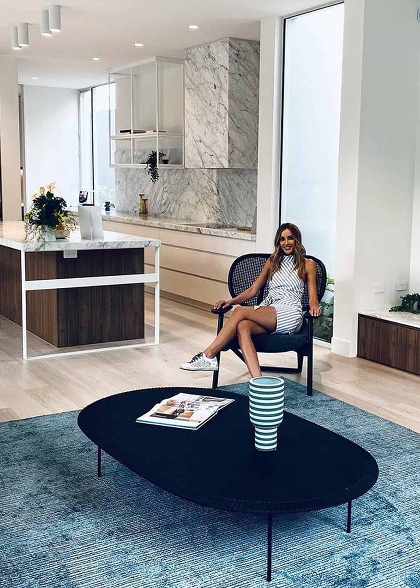 Woman sitting in an armchair in an open-plan living area with a dark oval coffee table and kitchen view.