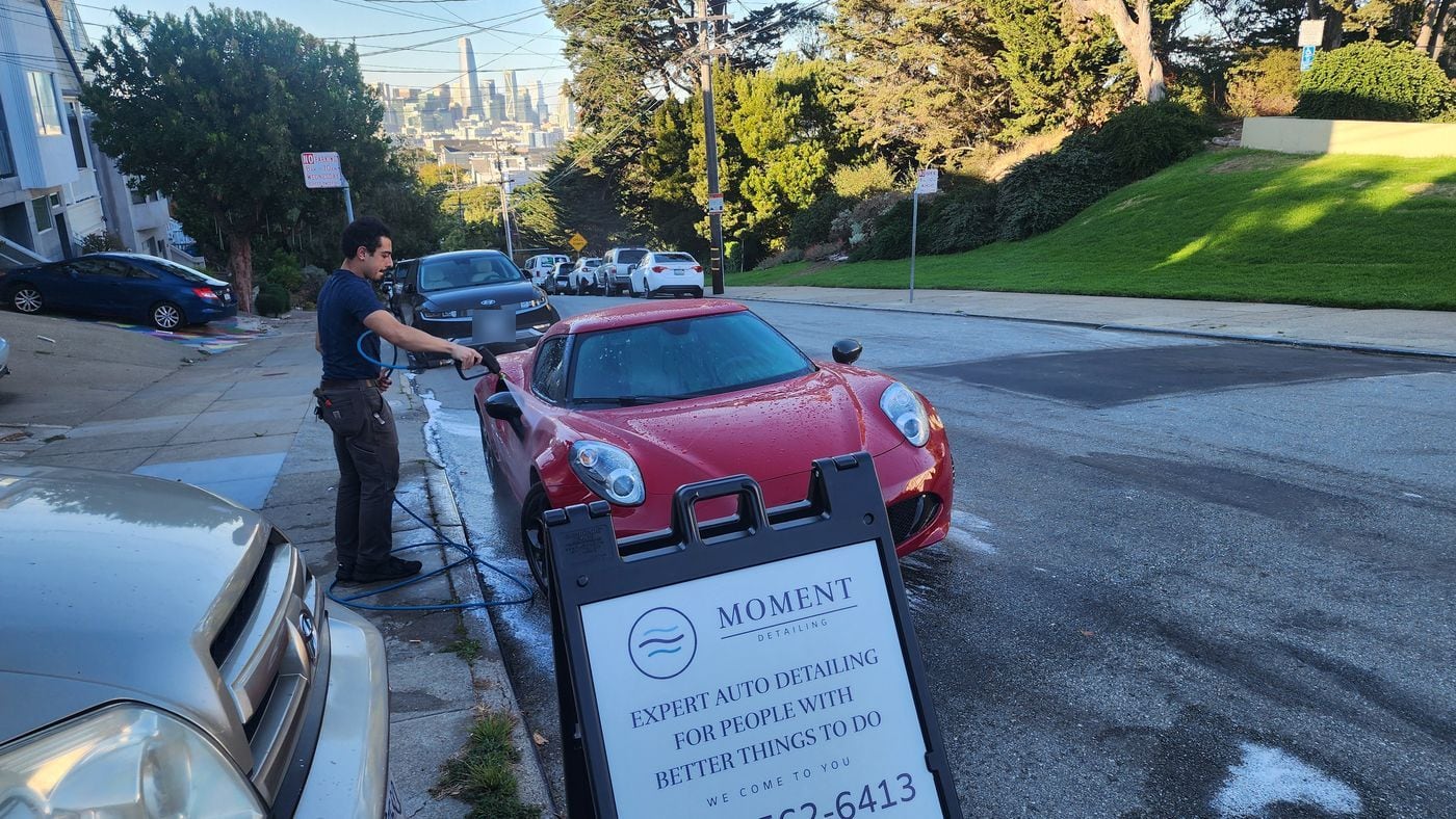 Home 12 Guillermo rinsing a red Alfa Romeo 4C beside his Moment sign, with the San Francisco skyline behind