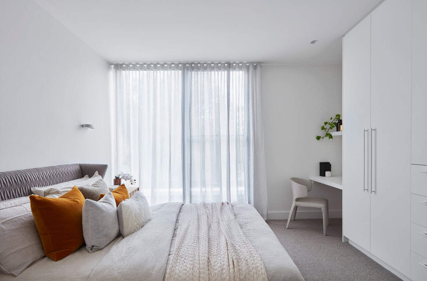 Bright modern bedroom with white wardrobes, study nook, mustard accent pillows, and sheer full-length curtains.
