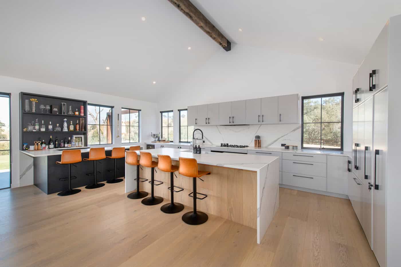 Open-plan kitchen and bar area with marble island, tan stools, dark liquor cabinet, and timber beam ceiling.