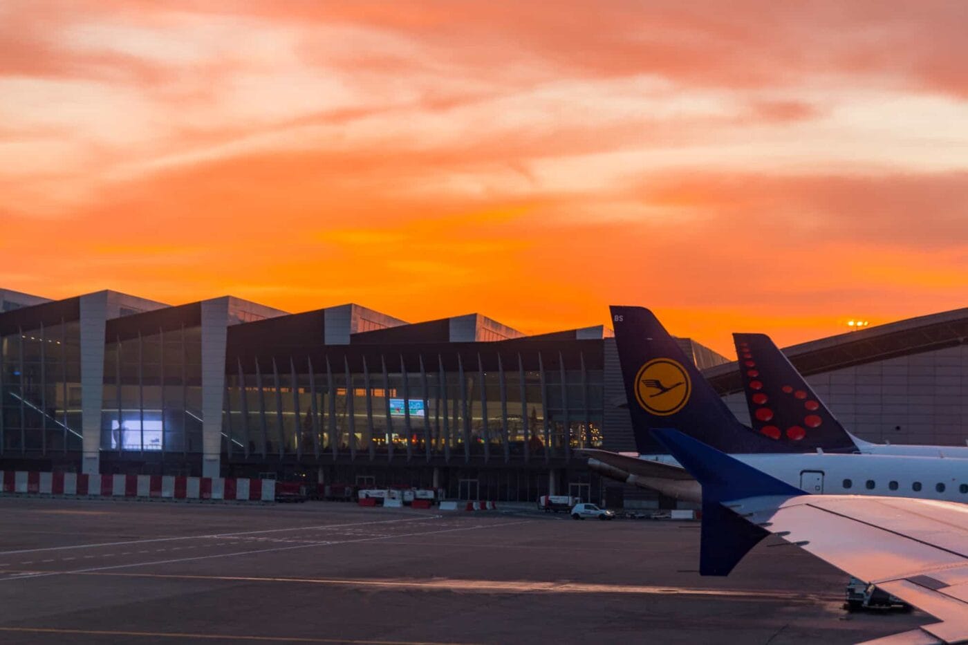 Looking out airplane window with wing in the photo and the airport in the background with bright orange sky.