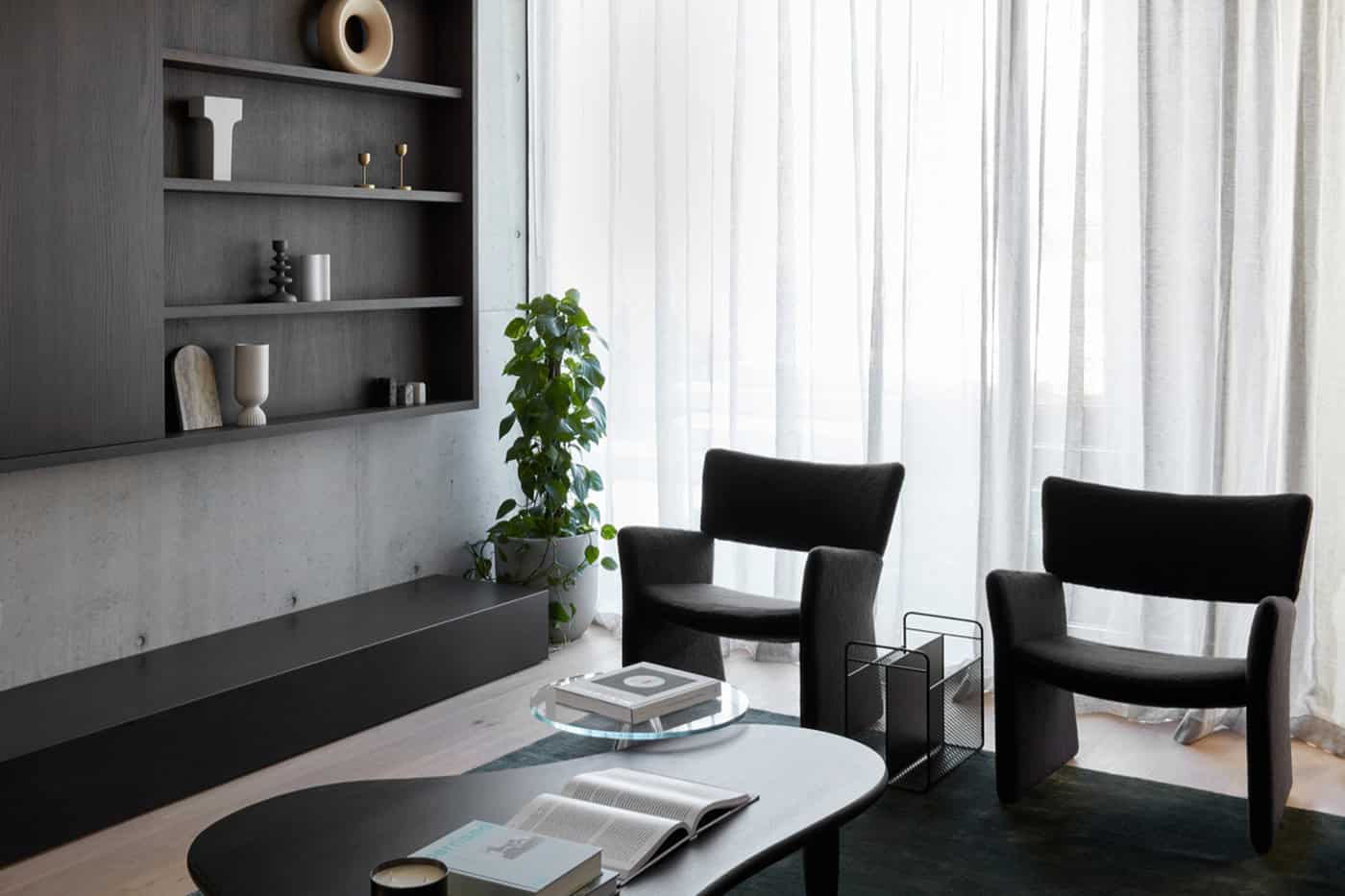 Pair of black armchairs beside sheer curtains and dark timber shelving in a modern living room.