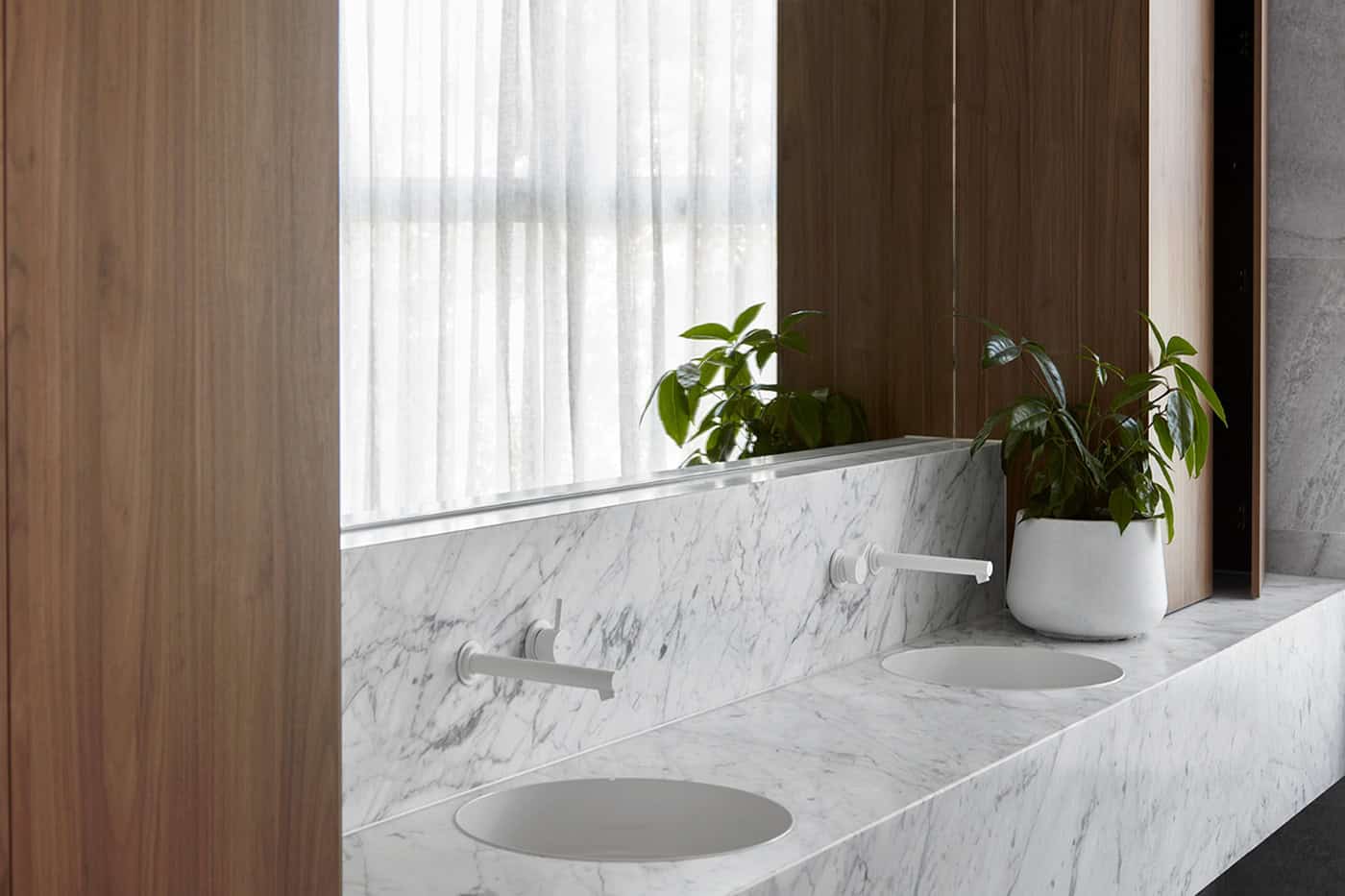 Close-up of a white marble vanity with dual undermount sinks, white wall-mounted taps, and a potted plant.