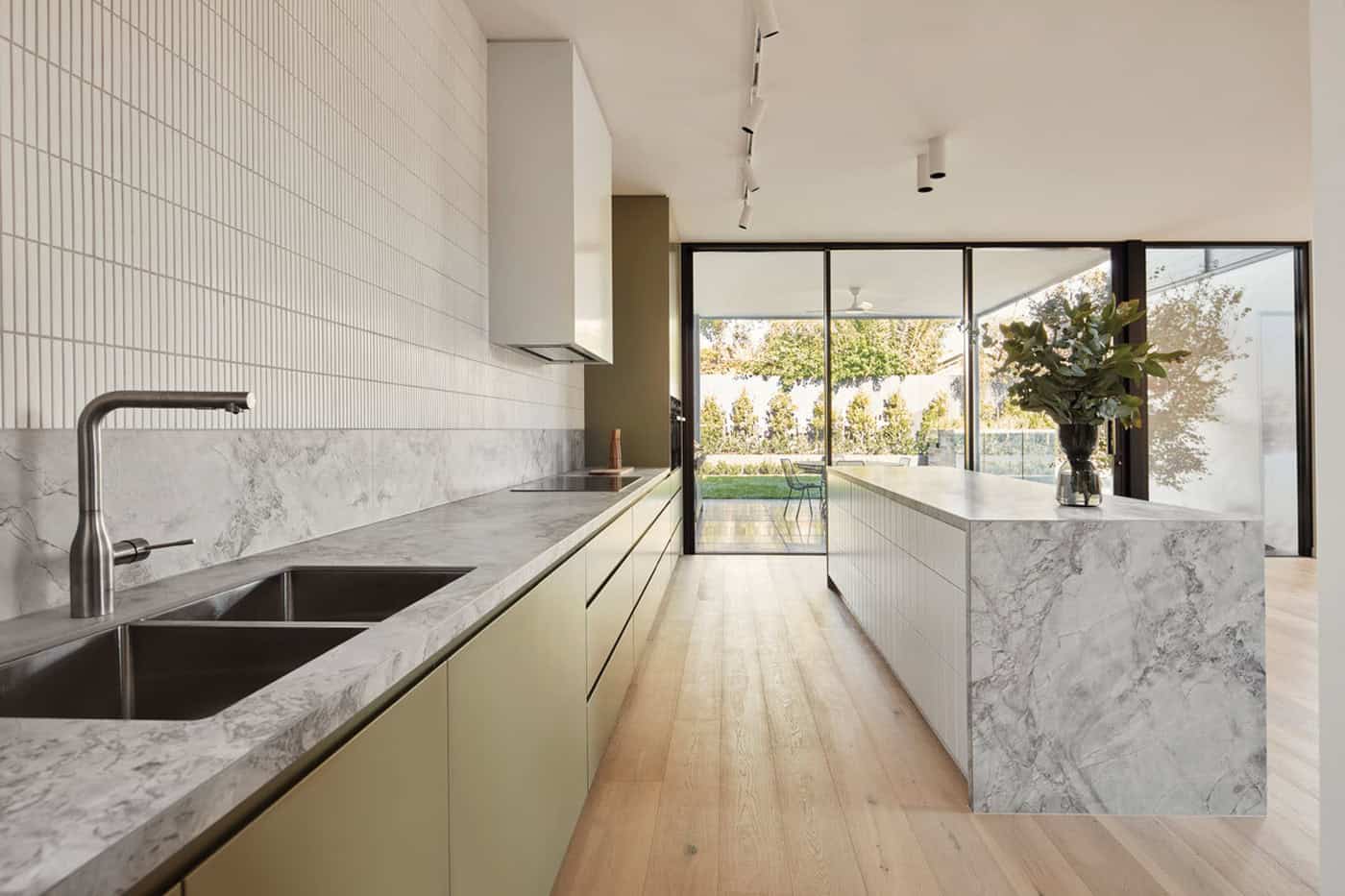Modern galley kitchen with marble countertops, green cabinets, kitkat wall tiles, and outdoor patio views.