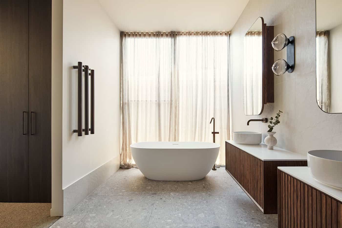 Luxurious bathroom with freestanding tub, fluted timber vanity, round sinks, dark fixtures, and sheer curtains.