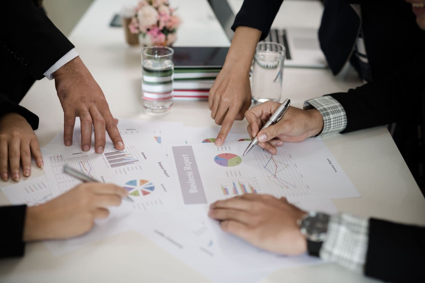 Foto aproximada de uma mesa de reunião, onde aparecem mãos escrevendo e folhas com gráficos em cima da mesa