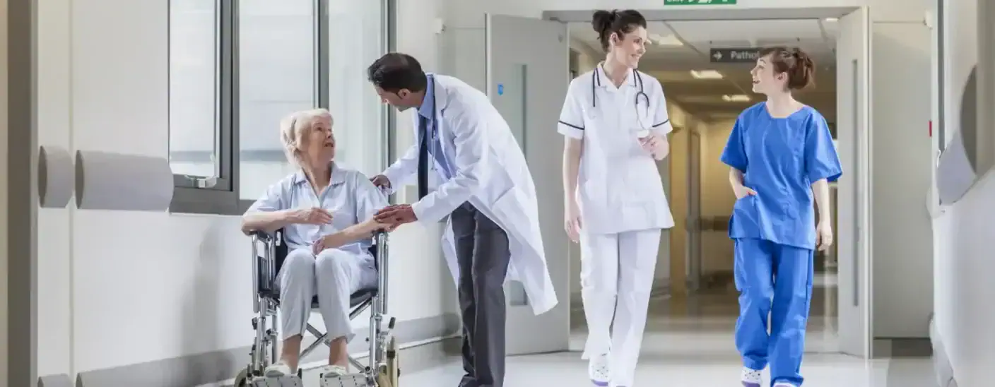 Doctor in healthcare sector assisting an elderly patient in a wheelchair while nurses walk through a hospital corridor