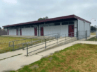 Single-story modular building with maroon doors and a flat maroon roof, accessible ramp with metal handrails leading to entrances on a grassy field under a cloudy sky.