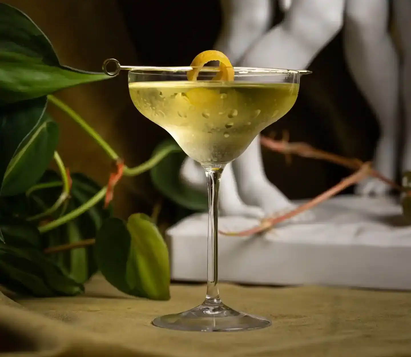Elegant craft cocktail served in a chilled coupe glass with a lemon twist garnish, styled with lush greenery and a classical sculpture for a refined Raleigh cocktail bar atmosphere in Midtown.
