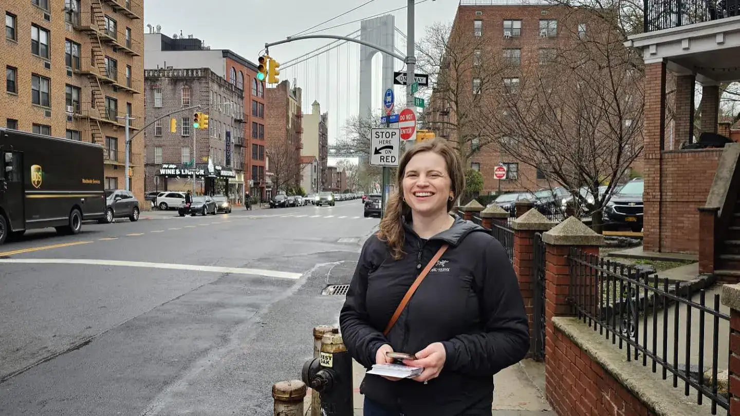 Kayla Santosuosso standing with campaign literature on 3rd Avenue in Bay Ridge, Brooklyn with the Verrazzano Narrows Bridge in the background.