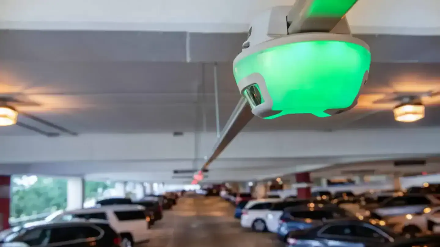 A close-up of a green M5 smart sensor, the heart of Park Assist's parking guidance system, installed on the ceiling of a parking garage