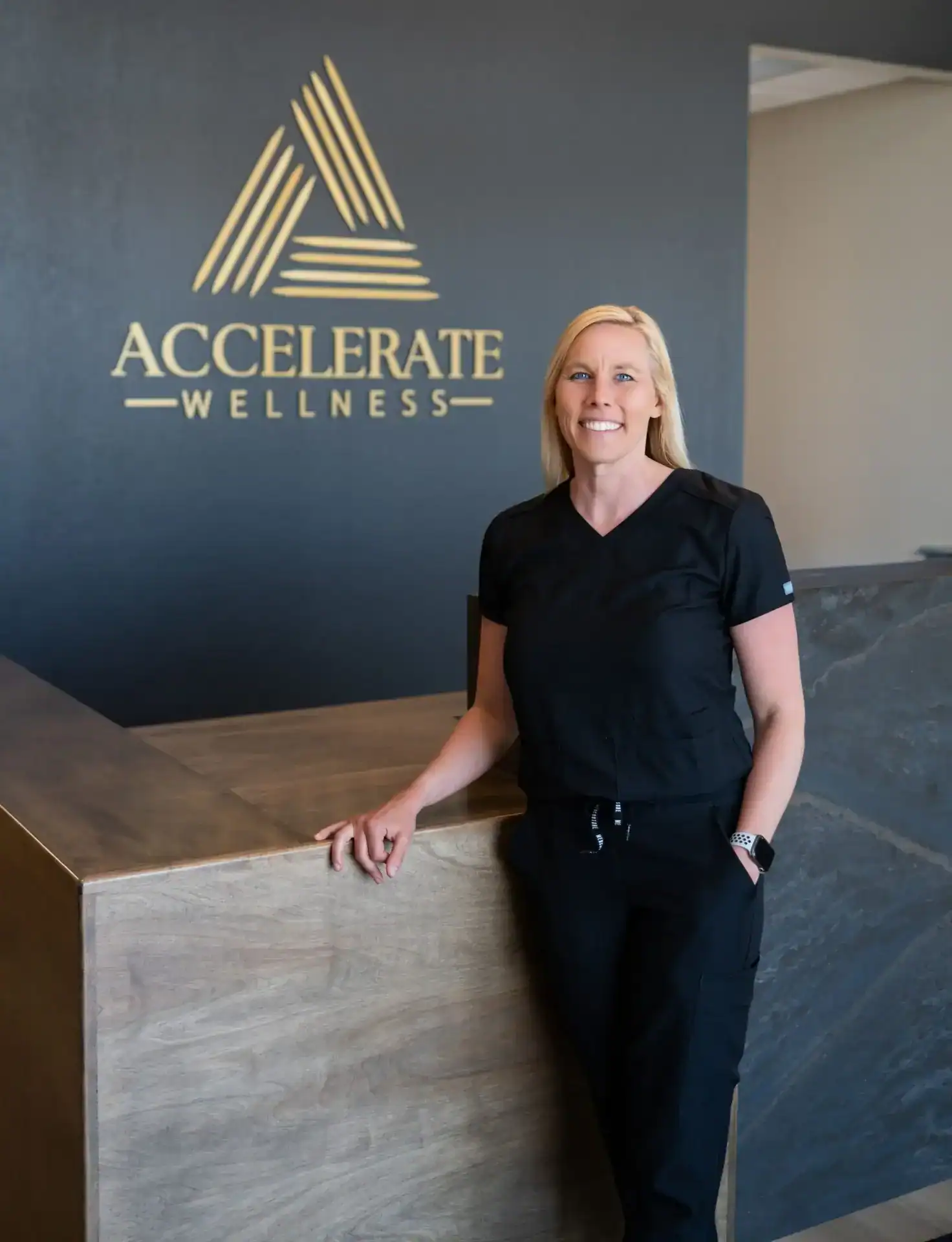 Kim Dewing, PA-C, smiling in black scrubs in front of the Accelerate Wellness logo wall.