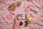 A high-end flat lay on pink fabric featuring a hand holding a smartphone with a self-care video, surrounded by chocolates, a latte, a rose, and floating holographic TikTok Gift Finder icons.