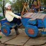 Фотография запечатлела маленькую девочку, сидящую на самодельной деревянной игрушке в форме автомобиля на игровой площадке, в окружении других детских игровых конструкций.  Композиция кадра построена таким образом, что главная героиня, девочка, находится в центре внимания, занимая правую половину изображения. Она сидит на ярко-синем деревянном игрушечном автомобиле, который занимает большую часть переднего плана. Автомобиль установлен на деревянной платформе, от которой отходят цепи, ведущие к пружинному механизму, что указывает на его качающуюся конструкцию. Взгляд девочки направлен прямо в камеру, что создает ощущение прямого контакта со зрителем. На заднем плане видны элементы более крупной детской площадки, выполненной из дерева, а также деревья и, вдалеке, мужчина, сидящий на скамейке, что дополняет атмосферу общественного пространства.  Ключевой субъект – девочка, возрастом примерно 7-8 лет, с длинными светлыми волосами, заплетенными в две косички. На голове у нее белая повязка с рисунком. Девочка одета в желтую ветровку, темно-синие штаны и коричневые детские кроссовки. Выражение ее лица спокойное, с легкой улыбкой, что передает ощущение уверенности и комфорта. Она сидит, держась одной рукой за борт игрушечного автомобиля. Игрушечный автомобиль, являющийся вторым важным элементом, сделан из грубо обработанных деревянных досок, окрашенных в ярко-синий цвет. Колеса автомобиля также деревянные, с красными акцентами. Конструкция выглядит самодельной, что придает ей особый шарм и аутентичность.  Этот снимок выполнен в жанре уличной фотографии, с акцентом на естественность момента. Техническое исполнение фотографии находится на высоком уровне: четкое изображение, правильная экспозиция и сбалансированный баланс белого. Стиль фотографии реалистичный, без излишней ретуши или художественных фильтров. Цветовая гамма снимка состоит из природных оттенков, с преобладанием синего, желтого и землистых тонов, что характерно для изображения уличной детской площадки. Креативный выбор заключается в фиксации повседневного момента из жизни ребенка, подчеркивая его радость и увлеченность игрой.  Сцена разворачивается на открытой игровой площадке, с песчаным покрытием, что типично для многих детских зон. Освещение в кадре естественное, дневное, рассеянное, что свидетельствует о пасмурном дне или съемке в тени деревьев. Это освещение мягко ложится на объекты, не создавая резких теней и подчеркивая текстуру дерева. На заднем плане видны зеленые кроны деревьев, что указывает на весенний или летний период. Общая атмосфера снимка – спокойствие, умиротворение и детская беззаботность. Окружающие элементы, такие как другие части игровой площадки и сидящий вдалеке мужчина, создают контекст для основной сцены, делая ее частью более широкого городского пейзажа.