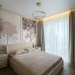 Интерьер светлой спальни в ЖК Level Причальный: кровать с бежевой обивкой, настенное панно с лесом, кольцевая люстра с хрусталем.