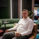 Портретная съемка с малой глубиной резкости. Денис Силавин в очках и светлой рубашке сидит вполоборота в мягком кресле лаунж-зоны, удерживая в руке стакан с кофе. Мгновенная фиксация расслабленной позы. На заднем плане размыты ритмичные вертикальные световые полосы зеленой стены стенда.