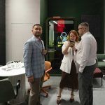 Репортажная фиксация цифрового взаимодействия. Рустем Мамин, Денис Медведев и сотрудница в белом жакете стоят плотной группой у кабины тишины и сосредоточенно изучают информацию на экране смартфона. Баланс теплого интерьерного света и глубокого зеленого цвета стен застройки.