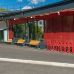 Крупный план уличных скамеек, прямоугольных урн и красной велопарковки на фоне витражного остекления спорткомплекса  