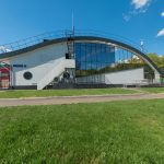 Современный фасад спортивного центра с масштабным витражным остеклением, ухоженным газоном и выровненной геометрией.