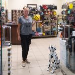 Молодая девушка со светлыми волосами, собранными сзади, и спокойным выражением лица стоит в полный рост в зоомагазине. В руках она держит и демонстрирует большую круглую белую клетку для птиц. Она одета в черную футболку с принтом и синие джинсы. На заднем плане расположена большая стена со встроенными вольерами для животных, а сверху на полках выставлены другие клетки разных цветов. Вверху видна яркая зеленая вывеска с надписью «КОШКИ».
